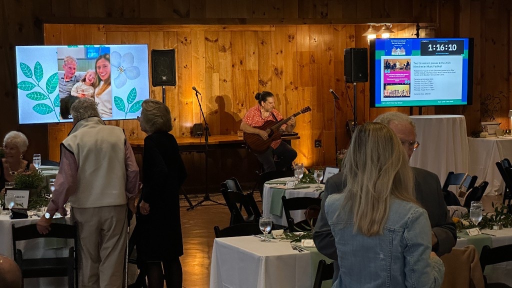 Live guitar in the barn, with student photos on one screen and an auction lot on the other.